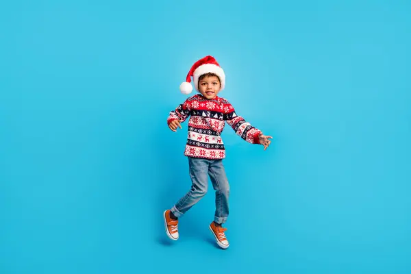 満足した子供のフルサイズの写真は 青い色の背景に隔離されたクリスマスパーティーで楽しんでジャンプ赤いプルオーバージーンズサンタ帽子を着用します — ストック写真