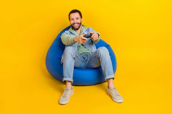年轻网游玩家手握无线操纵杆坐在豆袋上的全身照片 在黄色背景下被隔离得很有趣 — 图库照片