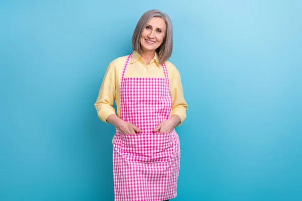 Portrait of pretty positive aged person toothy smile put hands apron pockets isolated on blue color background.
