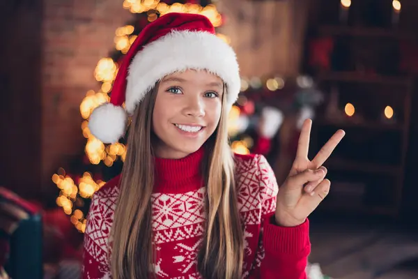 明るく陽気な小さな少女の写真は 家の部屋で新しい年を祝うV Signを示す赤いプリントセーターを着用しています — ストック写真