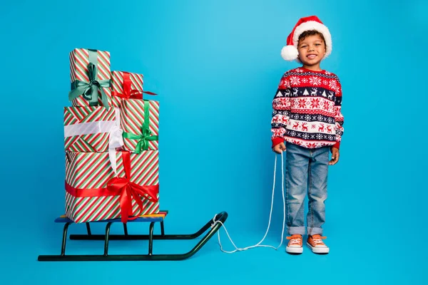 Full size photo of small little kid wear red pullover jeans santa hat hold sled with gift boxes by rope isolated on blue color background.