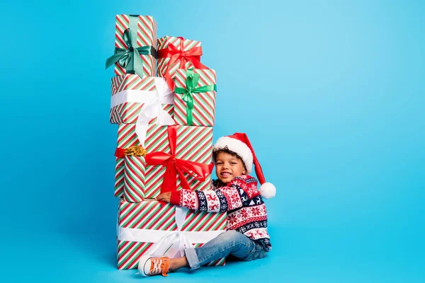 正面小孩身穿红色套头衫牛仔裤 头戴圣诞礼帽 坐在蓝色背景的礼品盒上的全副照片 — 图库照片