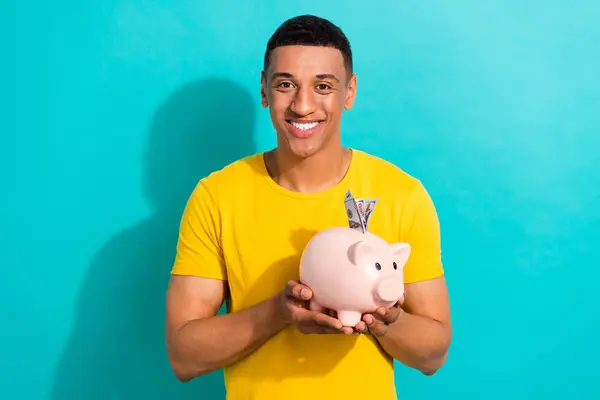 Photo of positive nice young man toothy smile arms hold money pig bank dollar banknotes isolated on teal color background.