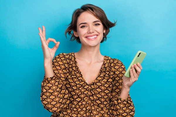 Photo of charming young woman bob brown hair show deal sign okay symbol enjoy smartphone facetime call isolated on blue color background.