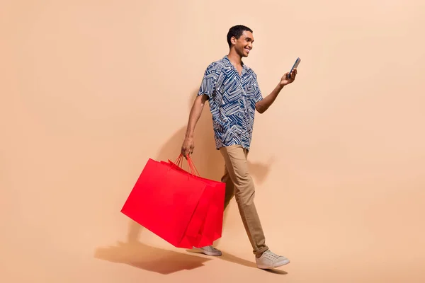 Full length photo of cool man dressed shirt hold shopping bags go empty space read email on smartphone isolated on beige color background.