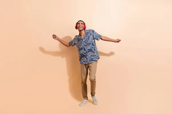 Full length photo of satisfied man dressed stylish shirt dancing in headphones look empty space isolated on beige color background.