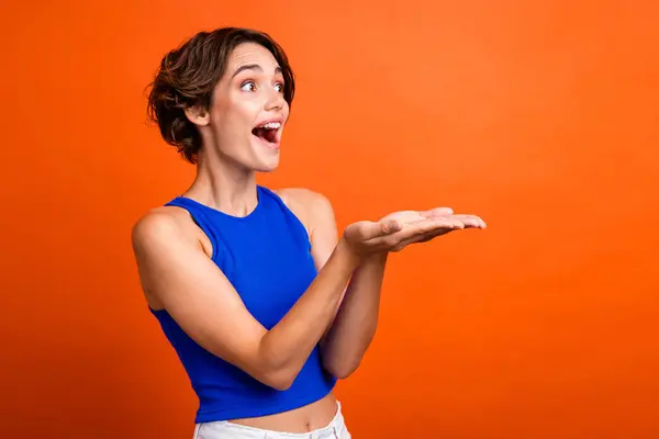 Photo of cheerful excited lady wear blue singlet open mouth looking showing arms empty space isolated orange color background.