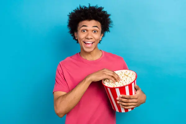 アフロ髪型の印象的な男の写真は ピンクのTシャツを着用し ブルーカラーの背景に隔離された映画でポップコーンウォッチ面白いフィルムを食べます — ストック写真
