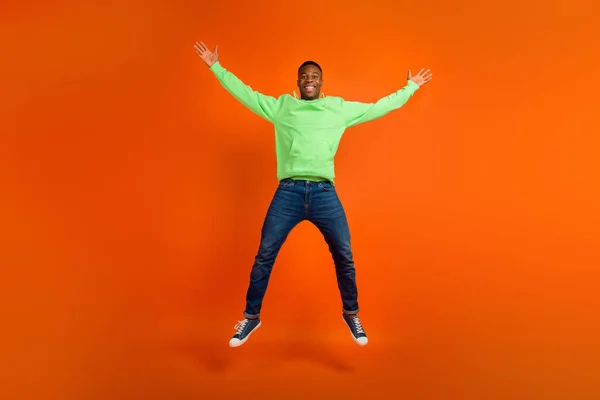 Full body photo of satisfied funky guy dressed green hoodie denim pants jumping demonstrate big sale isolated on vivid orange background.