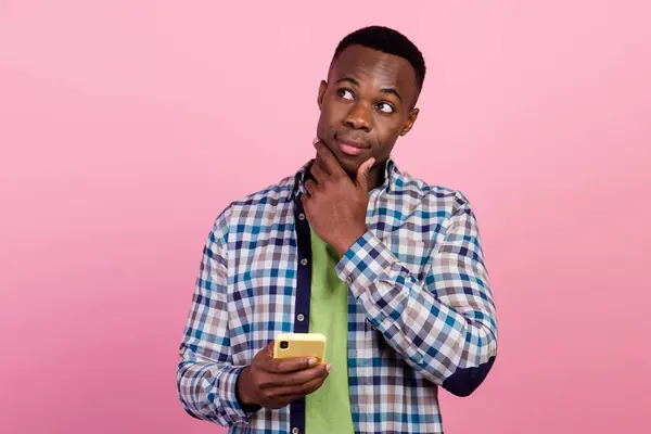 Photo of young content manager guy holding his smartphone thinking which video should post next isolated on pink color background.