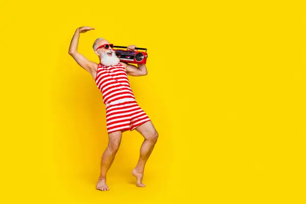 Full length photo of old man wear striped outfit dance tiptoes hold vintage music player look copyspace isolated on yellow color background.