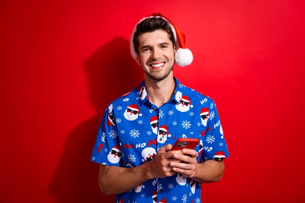 Portrait of clever friendly person dressed santa hat blue shirt holding smartphone read email isolated on vibrant red color background.