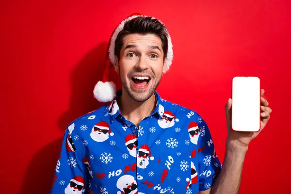 驚くほど興奮した男の写真クリスマスの帽子プリントシャツショー鮮やかな赤い背景で隔離されたスマートフォン画面の空のスペース — ストック写真
