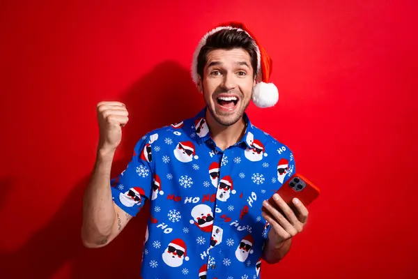 鮮やかな赤い色の背景で隔離されたスマートフォンを保持するクリスマスの帽子の印刷物のワイン宝くじを身に着ける熱狂的な男の写真 — ストック写真