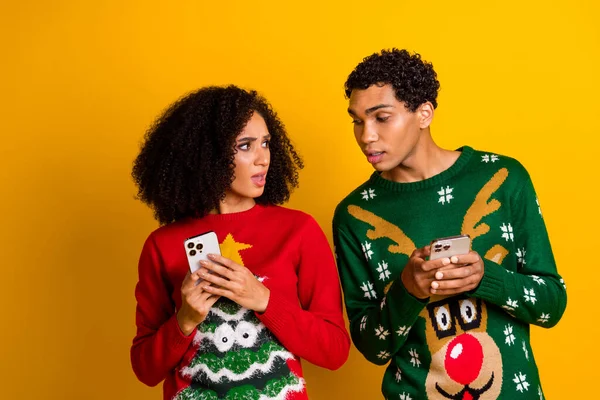 Photo of two people look curious hide smart phone texting message new year ugly sweater isolated on yellow color background.