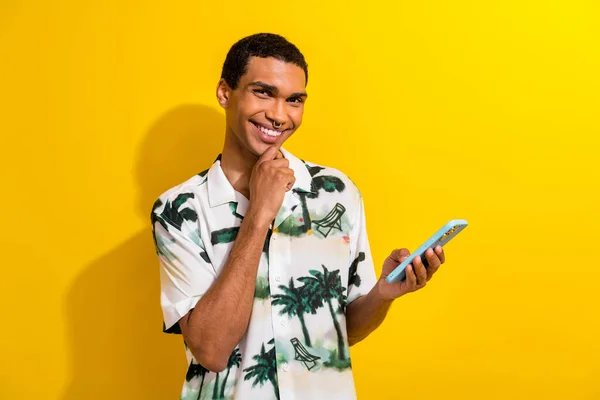 Portrait of smart positive guy with ring in nose wear print clothes hold smartphone hand on chin isolated on yellow color background.