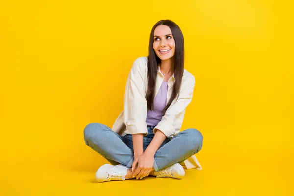 Full length photo of gorgeous positive minded person sit floor look empty space ad isolated on yellow color background.