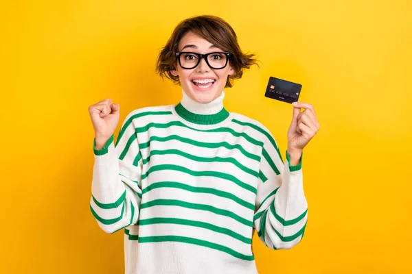 Photo of lucky excited lady wear striped pullover spectacles rising fist bank card isolated yellow color background.