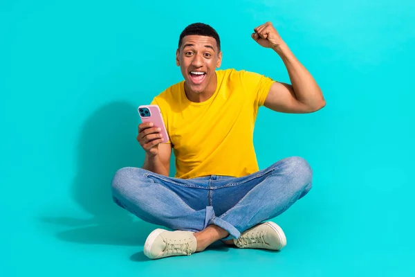 Full length portrait of positive funky man sit floor use smart phone raise fist success isolated on turquoise color background.