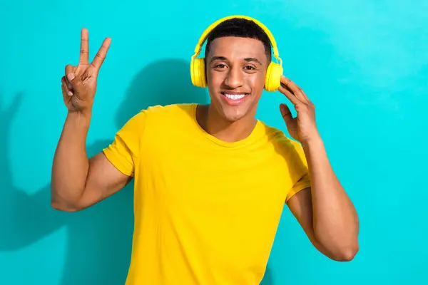 Photo of young happy guy wearing yellow t-shirt show v-sign hello advertisement modern headphones isolated on aquamarine color background.