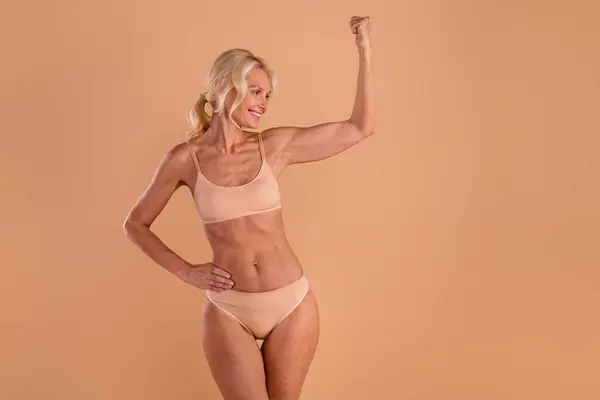 Photo of happy strong senior lady dressed lingerie showing arm muscles empty space isolated beige color background.