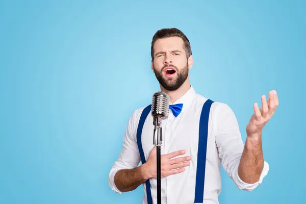 Portrait of talented attractive singer with bristle stubble in elegant classic outfit, singing hit with open mouth in microphone gesture with hand isolated on grey background.