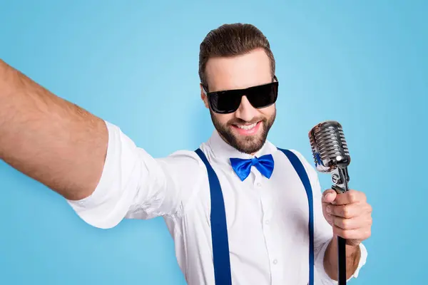 Self portrait of stylish elegant singer wearing bowtie shirt suspenders shooting selfie on front camera holding mic isolated on grey background.