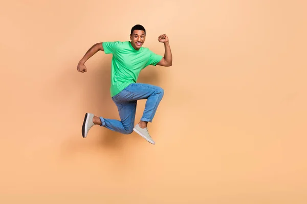 Full size photo of optimistic active guy dressed green t-shirt denim pants fast running in empty space isolated on beige color background.