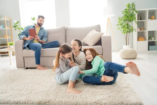 Full body portrait of four persons satisfied guy sit on sofa girls play lying on carpet toothy smile free time indoors.