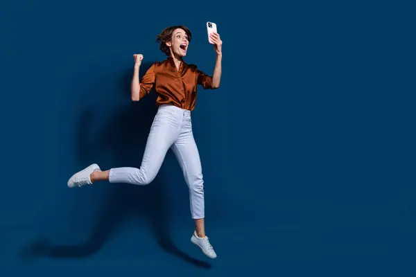 ラッキーファンキーな女性のフル長さの写真は ブラウンシャツジャンプゲームを着用しています 現代デバイス空のスペース ブルーカラーの背景 — ストック写真