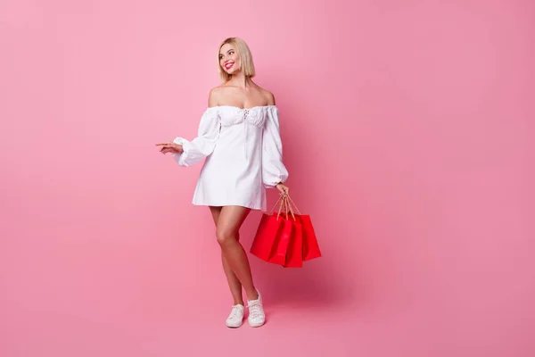 优雅可爱的女人穿着白色连衣裙的全长照片看着标识空旷的空间拿着用粉色背景隔开的购物袋 — 图库照片