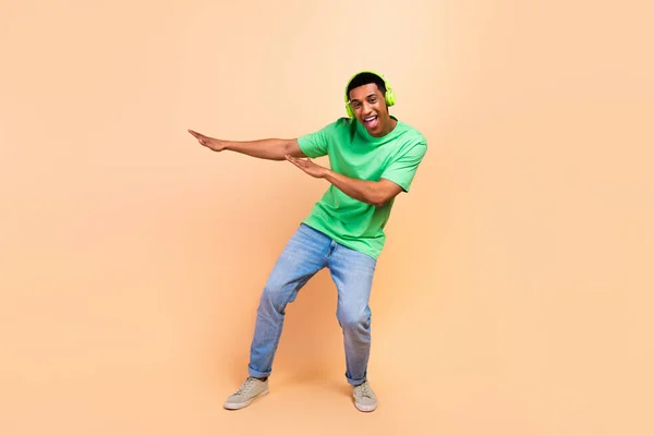 Full length photo of ecstatic positive man wear stylish t-shirt jeans listen musin in headphones dance isolated on beige color background.