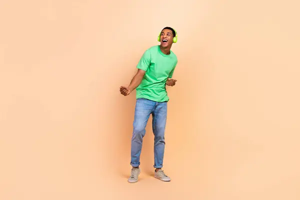 Full length photo of positive man wear stylish t-shirt jeans in headphones dancing look empty space isolated on beige color background.
