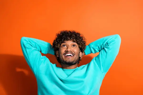 Portrait of minded creative person toothy smile arms behind head look up empty space isolated on orange color background.