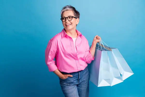 Portrait of funny person wear stylish blouse in eyewear hold shopping bags look at promotion empty space isolated on blue color background.