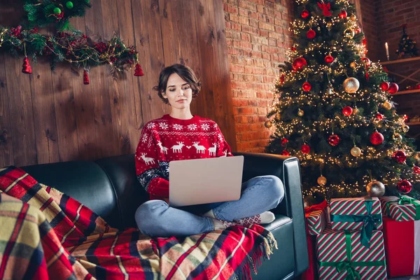 坐在皮革沙发上可爱的女孩实现梦想的全身像笔记本电脑在家中遥不可及的室内装饰圣诞树旁 — 图库照片