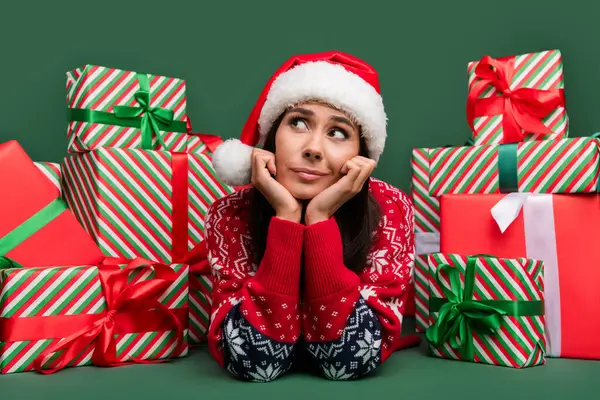 Portrait of funny thoughtful young girl santa claus helper touch cheeks look minded gift boxes new year isolated on green color background.