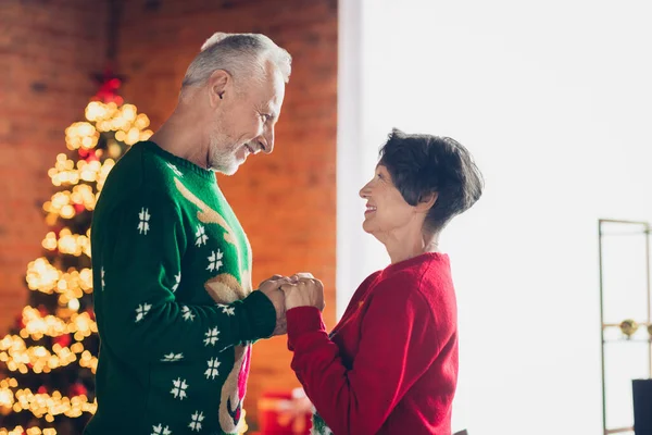 Side profile photo of old couple pensioners hands together look each other smiling love story all life xmas spirit wear funny ugly sweaters indoors.