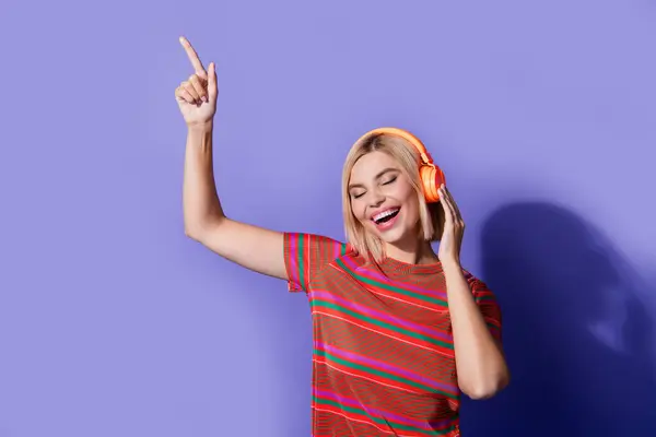 紫色の背景に隔離されたオレンジ色のヘッドフォンに残されたストライプTシャツポイントフィンガーの写真 — ストック写真