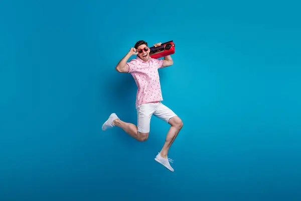 Full size photo of nice sporty person jumping carry boombox arm touch sunglass isolated on blue color background.