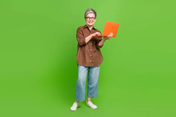 Full length photo of smart clever woman dressed oversize shirt jeans hold laptop typing message isolated on green color background.