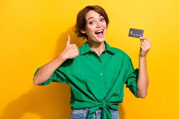 Photo of funky excited lady wear green shirt showing thumb up holding bank card isolated yellow color background.
