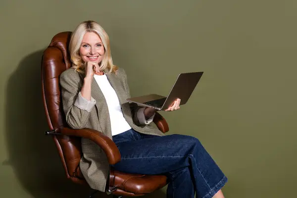 坐在棕色扶手椅上的快乐成熟的商务女士微笑的照片在卡其色背景上保持无线计算机隔离 — 图库照片