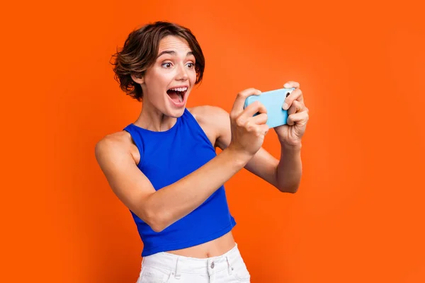 Photo of positive funky nice girl open mouth hands hold smart phone play games isolated on orange color background.