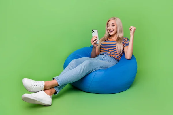 Full length photo of happy youth girl enjoy jackpot win hold smartphone sitting bean bag entertainment isolated on green color background.