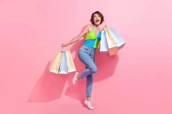 多くのショッピングバッグを持っているケアフリーファッショニスタの若い面白い少女のフル長さの写真は ピンク色の背景に隔離された空のスペースに見えます — ストック写真