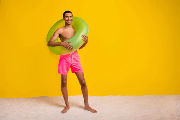 Full length photo of good mood handsome shirtless man walking beach ready swim floating ring empty space isolated yellow color background.