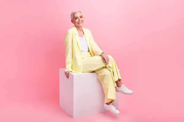 スタイリッシュなポジティブな快適な女性のフルサイズの写真は ピンク色の背景に隔離された白い立方体に座って黄色のスーツを着用しました — ストック写真