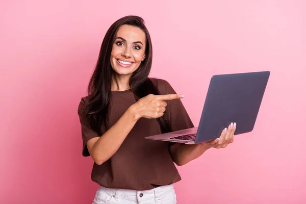 Photo of young woman in brown t shirt makes presentation point finger her new retina display laptop isolated on pink color background.