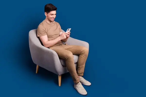 Portrait of intelligent man with stubble wear beige outfit sit on armchair typing sms on smartphone isolated on dark blue color background.